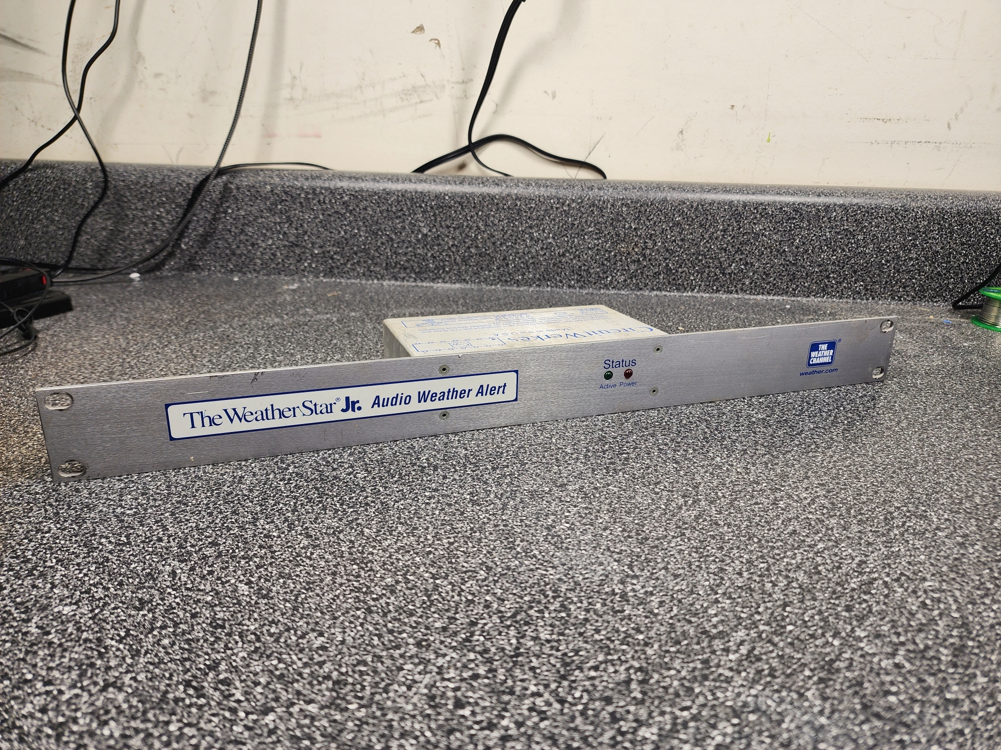 Picture 1: The WeatherStar Jr Audio Alert. It's a metal 1U chassis with a blue-outlined label on the front, reading: 'The WeatherStar Jr. Audio Weather Alert.' In the middle of the chassis, there is text that reads 'Status,' underneath it are two LEDS: one green, a label underneath reading 'Active', and one red, underneath is a label reading 'Power.' To the far right of the unit there is a blue logo for The Weather Channel, with 'weather.com' underneath.