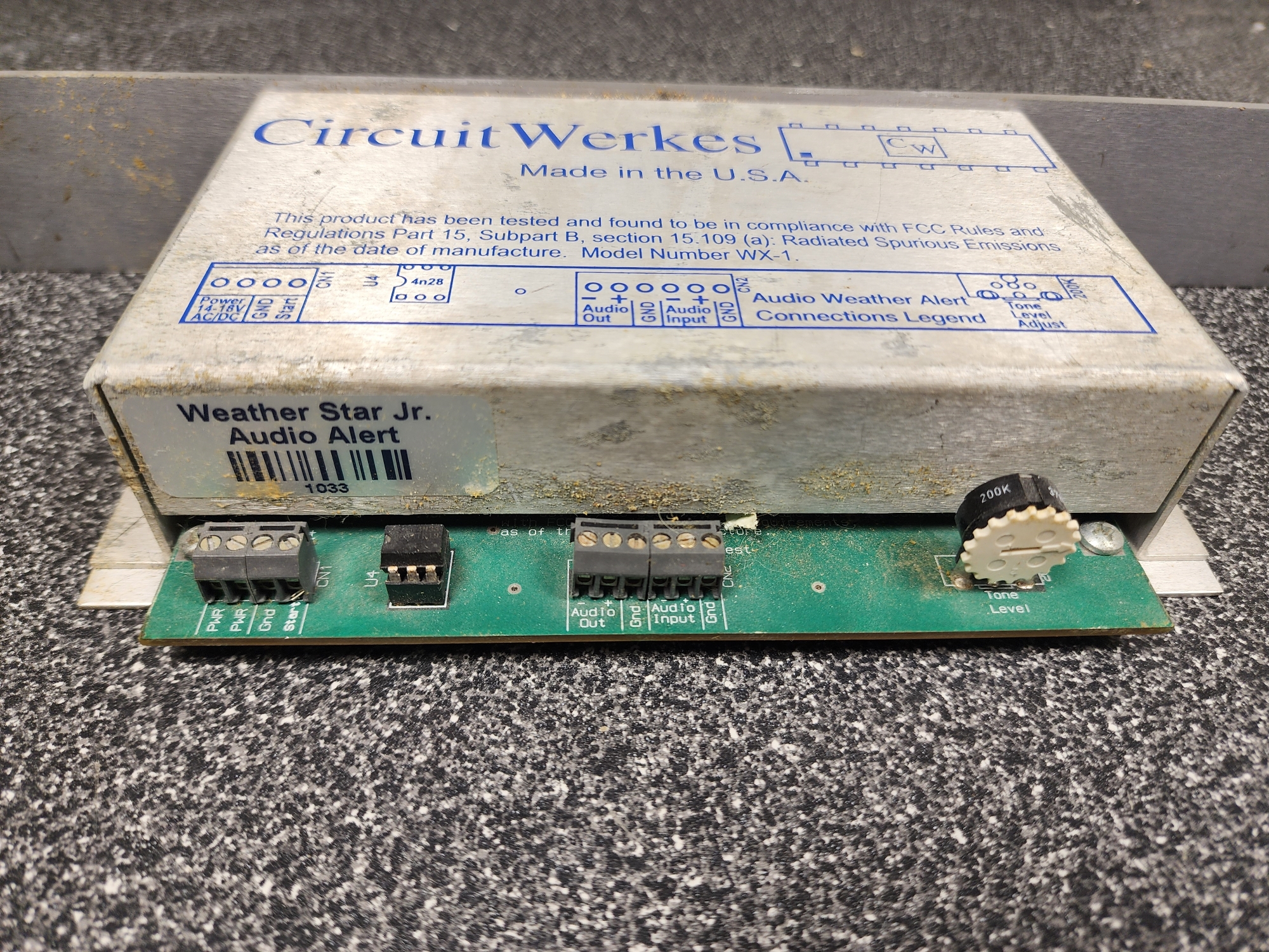 Picture 3: The rear side of the Audio Alert, with a bar code sticker numbered 1033. A green circuit board is sticking out of the metal enclosure, presenting 4 terminal blocks, a socketed chip, and a potentiometer.