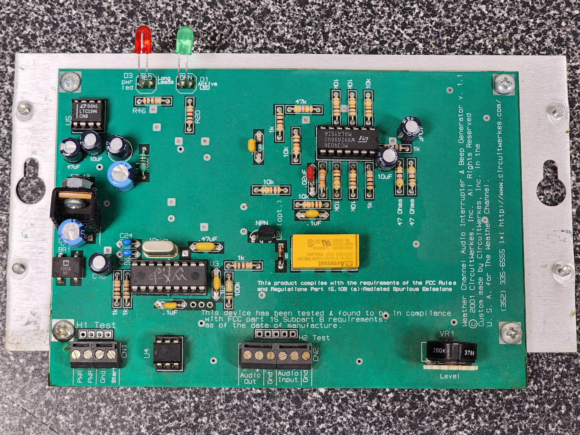 Picture 4: A top-down view of the Audio Alert circuit board. At the top left there is a red and green LED. There are several components on the motherboard. There is legible text in the middle near the bottom, which reads: 'This product complies with the requirements of the FCC Rules and Regulations Part 15.109 (a): Radiated Spurious Emissions,' a blurb below that reads: 'This device has been tested and found to be in compliance with FCC part 15 Subpart 8 requirements as of the date of manufacture.' On the right, there is an angled blurb reading: 'Weather Channel Audio Interrupter & Beep Generator V.1.1 Copyright Symbol 2001 CircuitWerkes, Inc. All Rights Reserved. Custom made by CircuitWerkes, Inc. in the U.S.A. for The Weather Channel. (352) 335-6555 http://www.circuitwerkes.com'